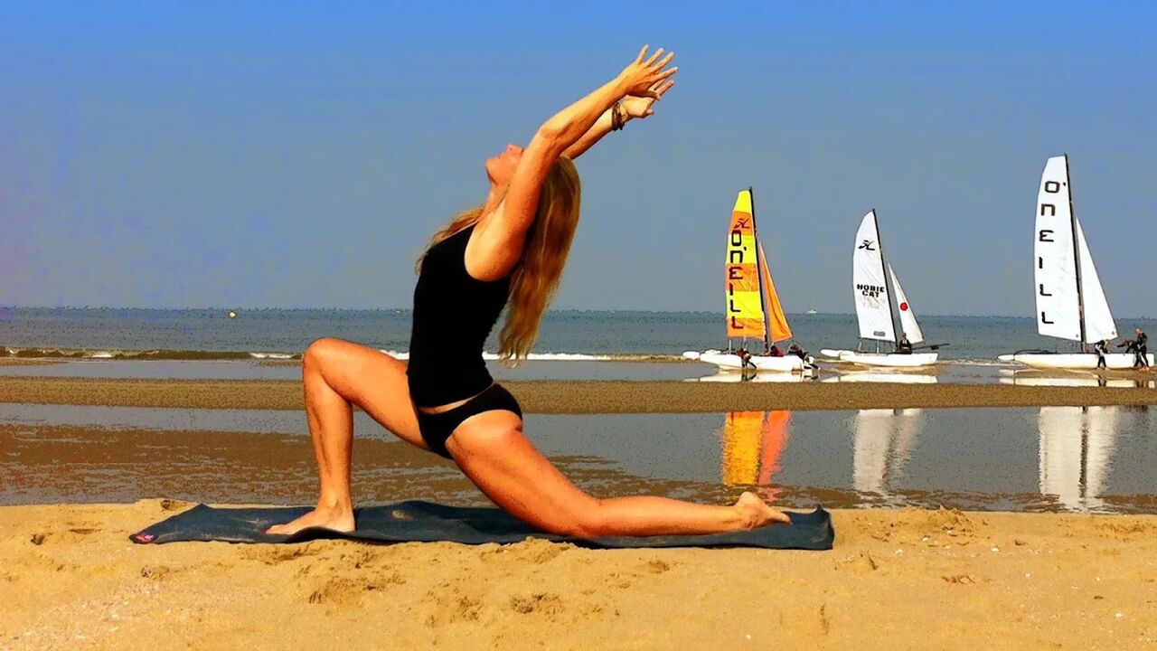Caroline van den Ende in een lage lunge met de armen omhoog op het strand, met catamarans op de achtergrond